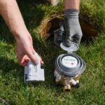 A person knealing on green grass shows updated meter equipment next to an irrigation hole.