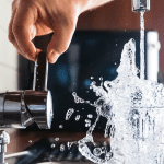 A hand holds the edge of a water faucet while water splashes into a water glass.