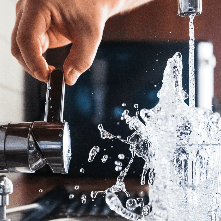 A hand holds the edge of a water faucet while water splashes into a water glass.