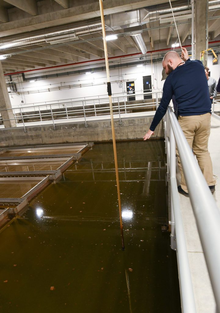 A bald white man lowers a yellow pole into a water treatment pool to test water quality.