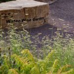 A tan stone bench labeled City of Thornton, Water-Wise garden sits in crushed gravel surrounded by natural grasses and shrubs.