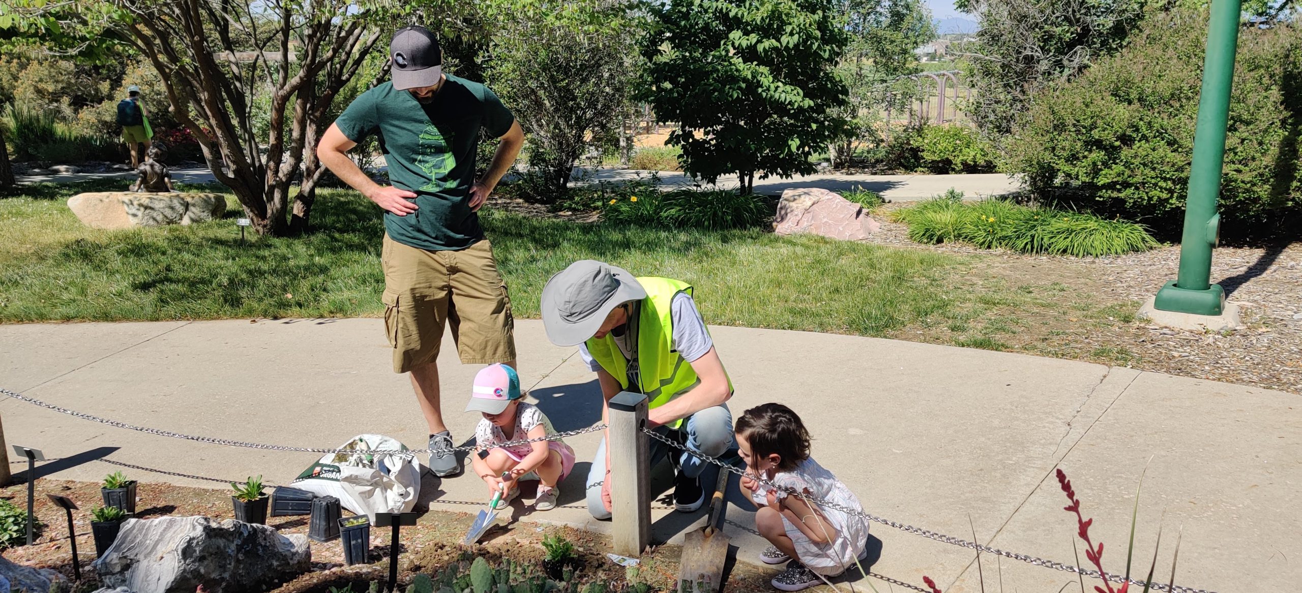 Man in bright green vest and light grey hat helps two children with shoveling into dirt. They are on a garden pathway with a man in a green shirt standing behind them.