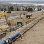 Dirt construction site with two yellow excavators digging into the ground. On the left there is a two-lane road.