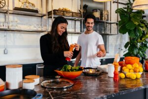 Woman and man with dark hair cook in a kitchen with open shelving and red bowl.