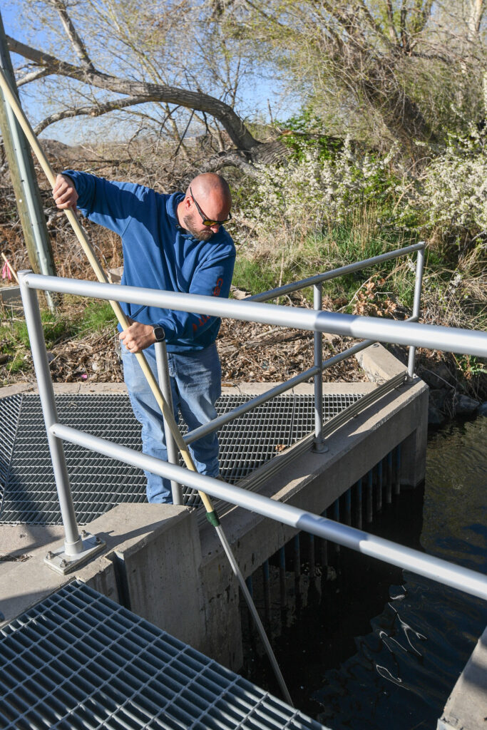 Man stands on metal grate over a ditch, holding long metal pole.
