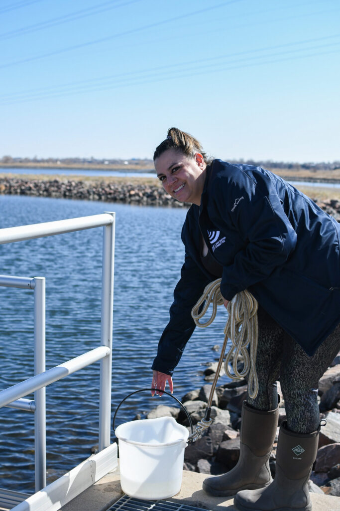 Person with dark blue jacket, grey leggings and waterproof boots holding a rope and bucket a platform over a Colorado reservoir.