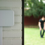 White irrigation controller box on a white brick wall on the left with a glowing blue icon. On the right, a blurry shot of a man playing with a large brown dog on grass.