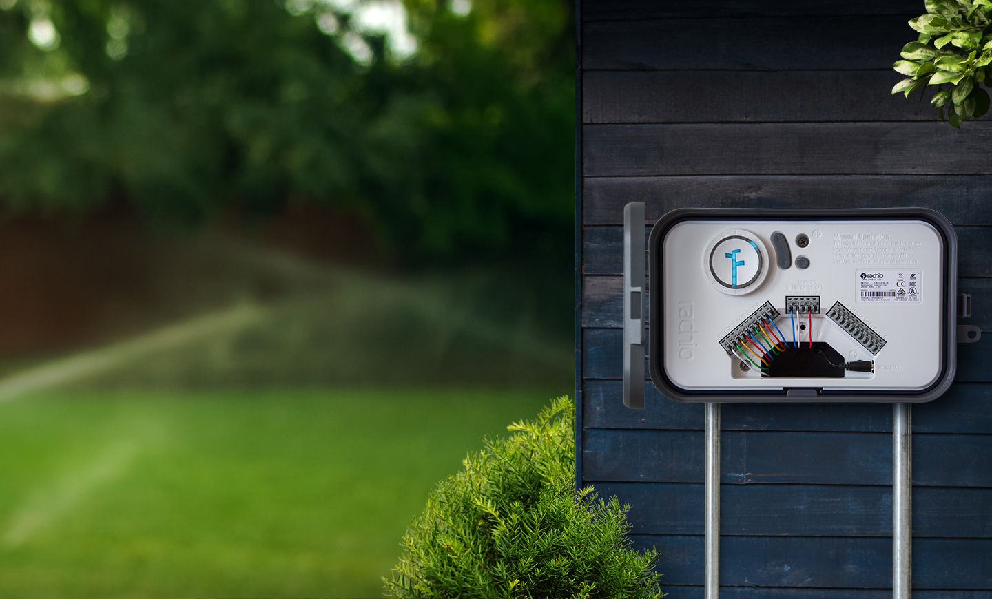 rachio-controller-with-outdoor-enclosure White irrigation controller box on a black wall with wires next to a blurred lush green lawn and trees.