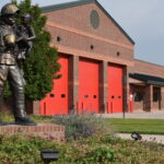 Metal statue of a firefighter holding a young child surrounded by lush green plants and a brick fire station with red doors on the right.