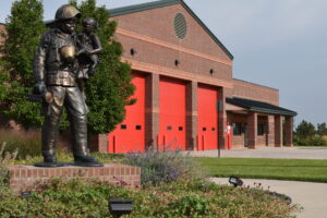 dsc_4445 Metal statue of a firefighter holding a young child surrounded by lush green plants and a brick fire station with red doors on the right.