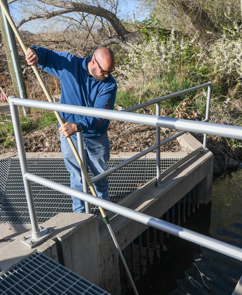 supply-10 A man in a blue sweatshirt uses a long pole to clear debris from a water infrastructure ditch.