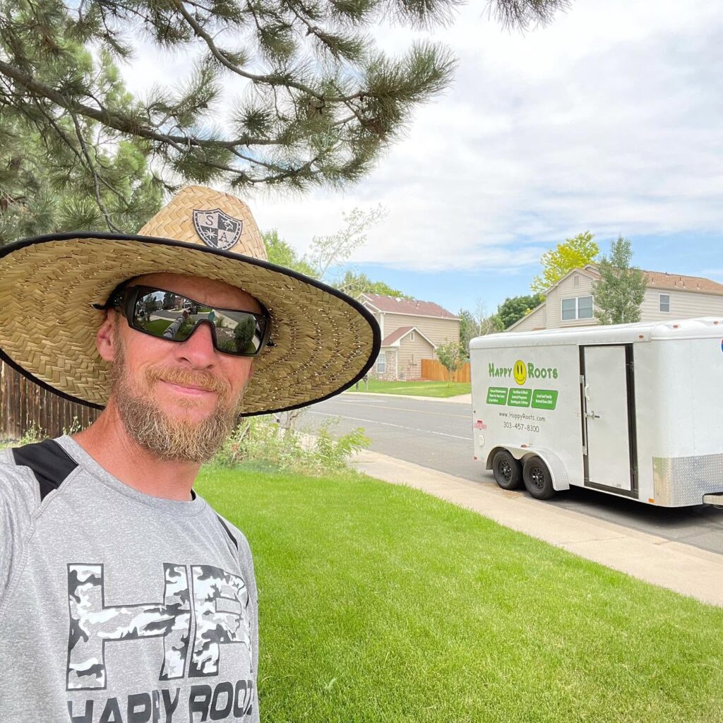 les-with-happy-roots2 A bearded man in sunglasses wearing a straw hat takes a selfie in front of a manicured lawn. A trailer with the words "Happy Roots" is visible in the background